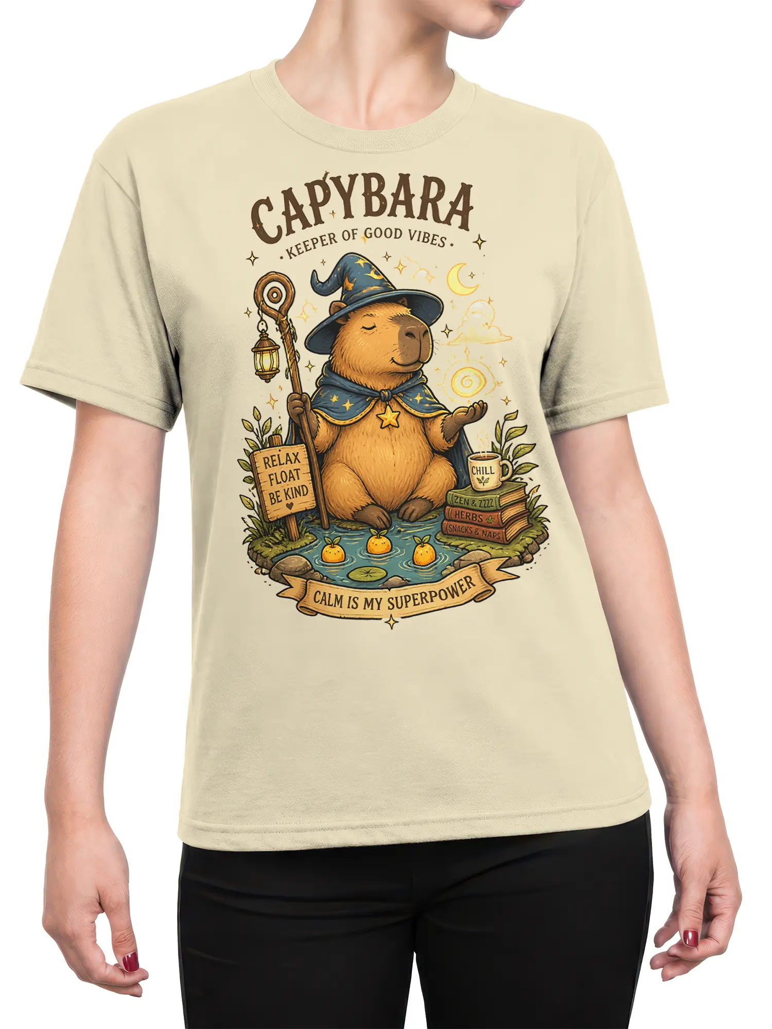 Female torso wearing a beige wizard capybara T-shirt with a cozy fantasy pond scene and calm is my superpower artwork