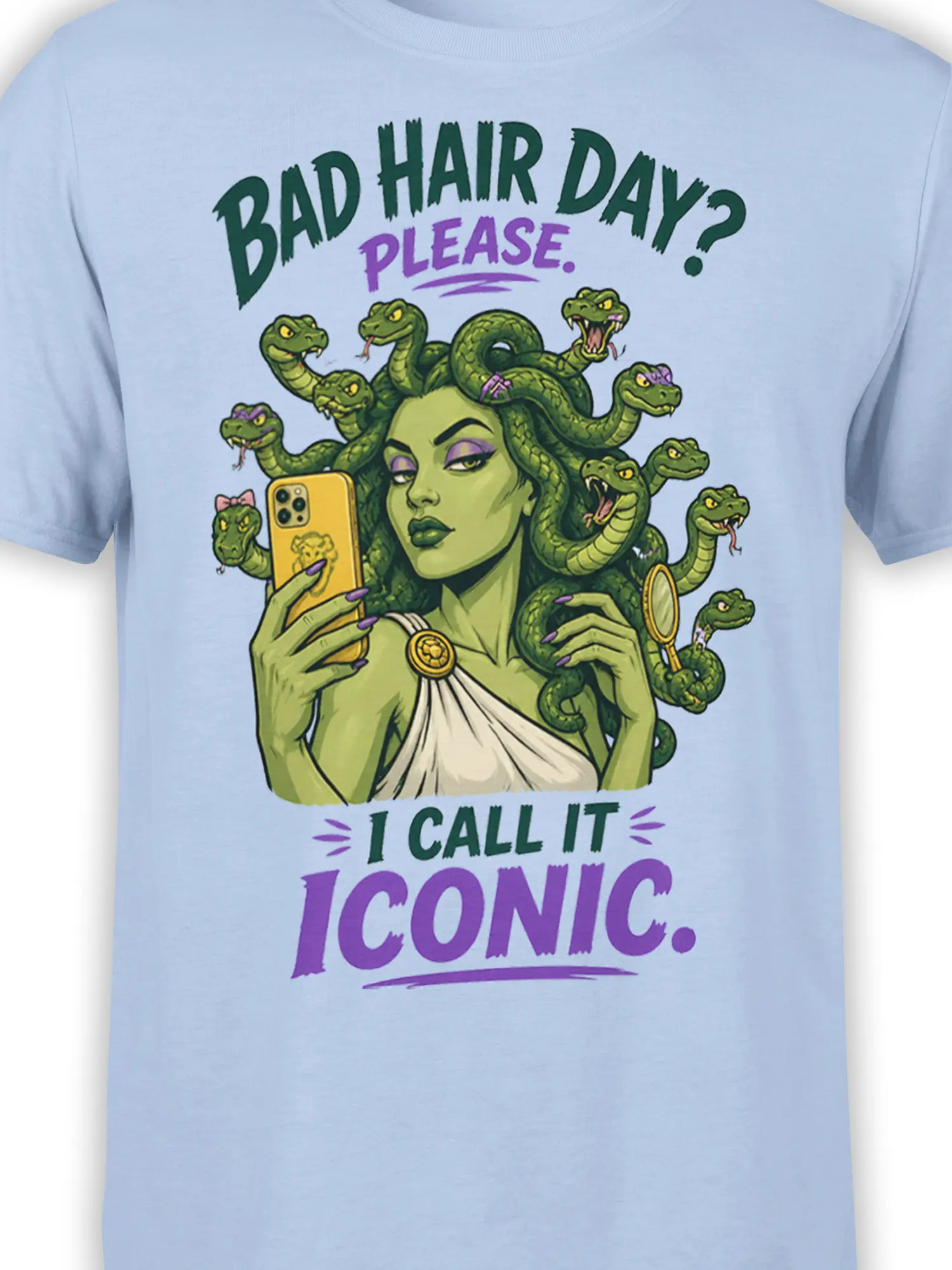 Close-up of green Medusa artwork with snake hair, purple details, phone, mirror, and bold text on a light blue shirt